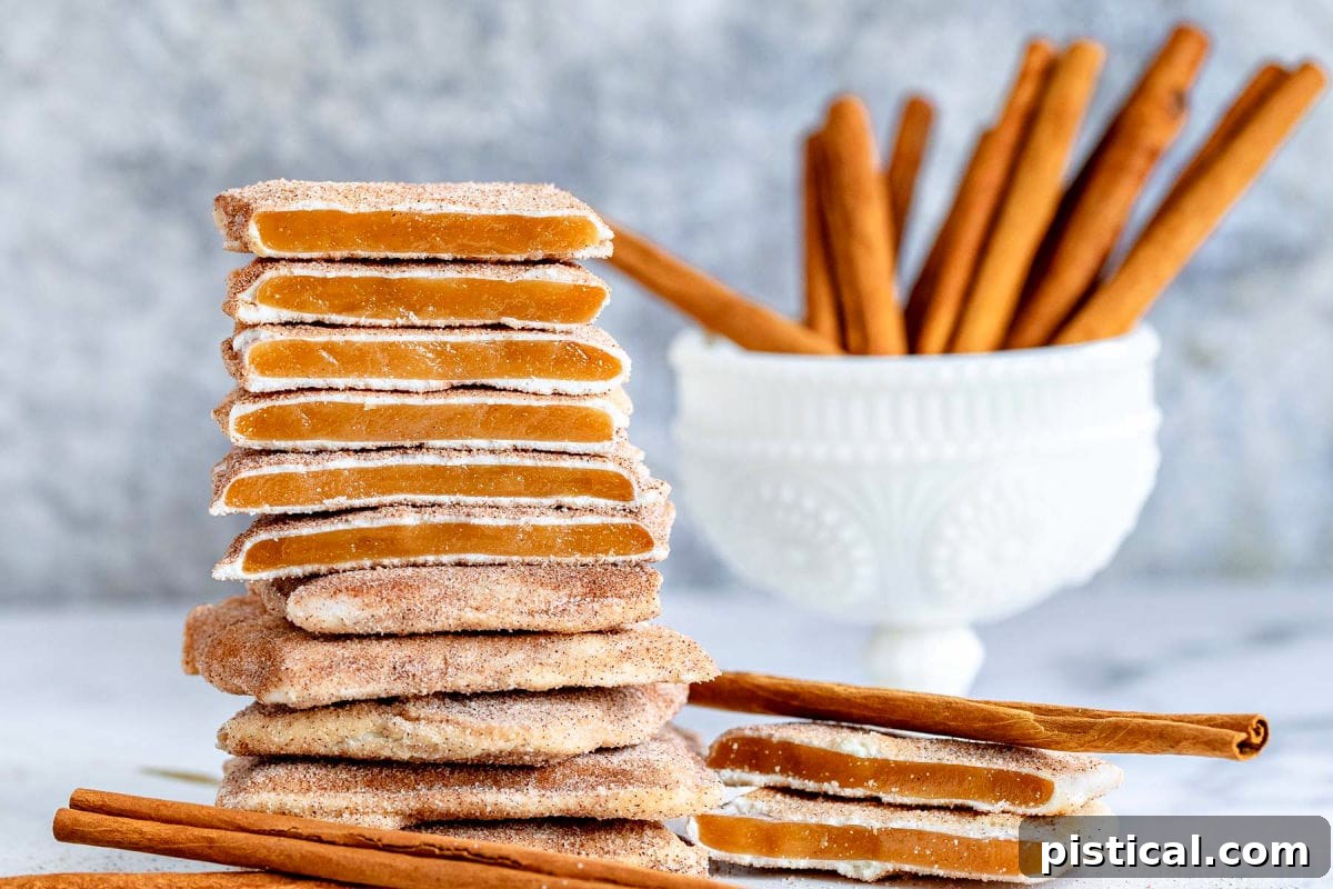 Close-up shot of square Churro Toffee pieces stacked, showing the distinct layers of toffee, white chocolate, and cinnamon sugar, with cinnamon sticks in the background.