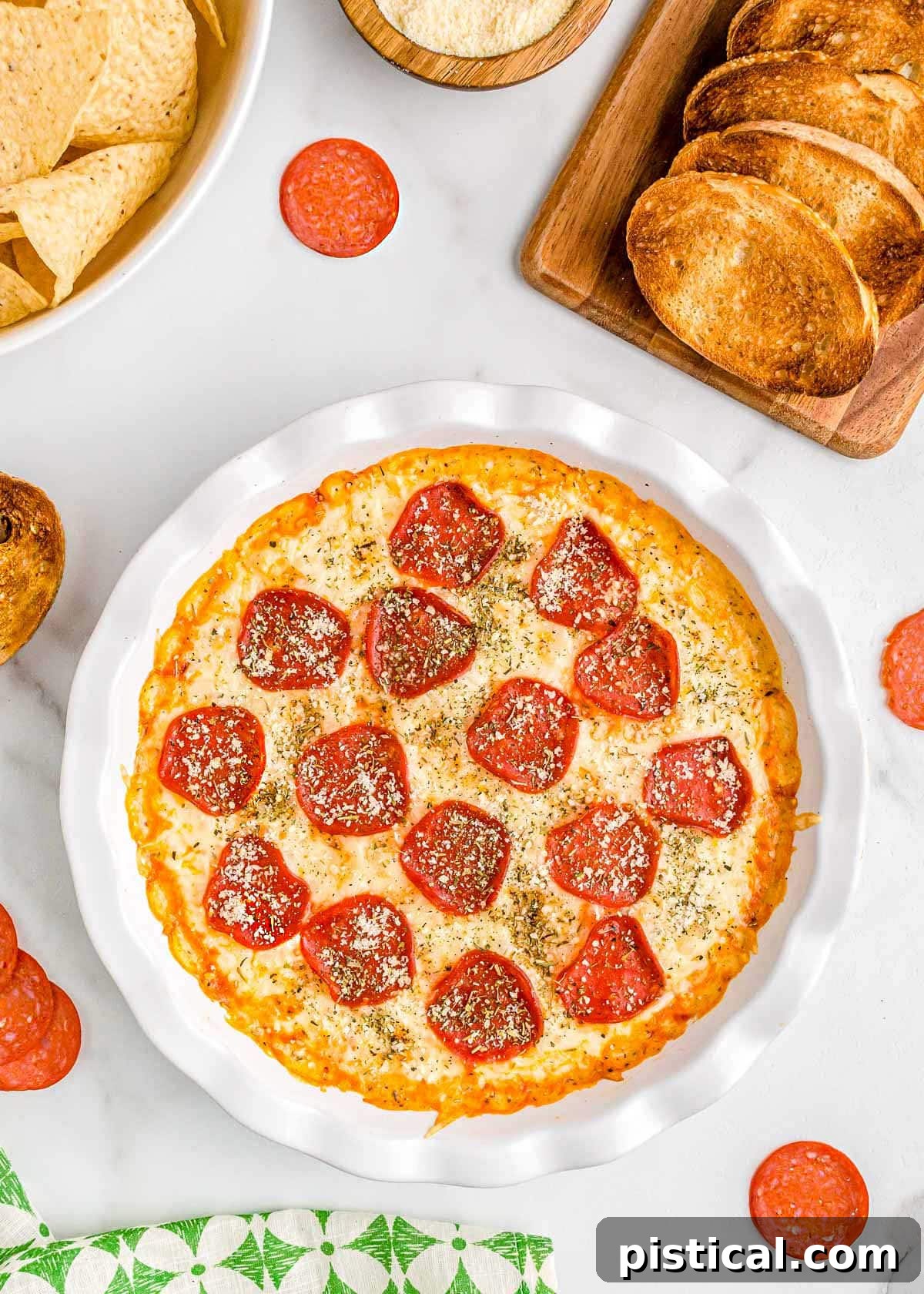 Ultimate Baked Pizza Dip 2 pizza dip baked in white pie dish topped with pepperoni and parmesan cheese. plate of toasted baguette and a bowl of chips are set off to the side.