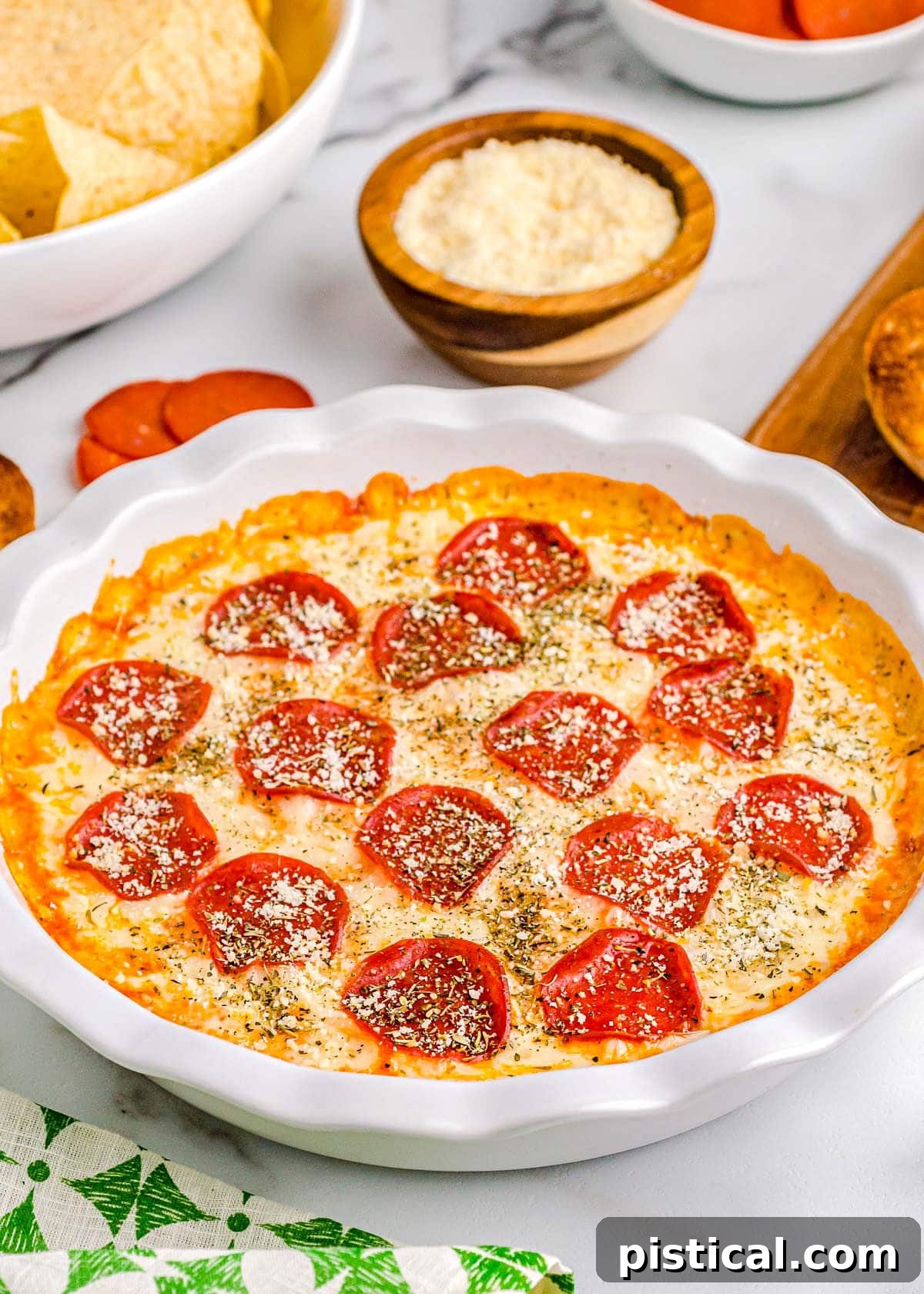 Ultimate Baked Pizza Dip 6 pizza dip baked in white pie dish topped with pepperoni and parmesan cheese. plate of toasted baguette and a bowl of chips are set off to the side.