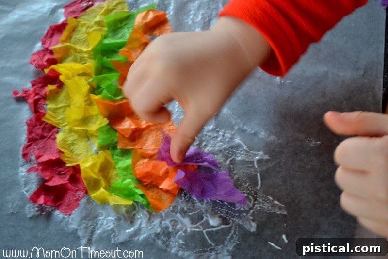 Child arranging colorful torn tissue paper pieces onto glue-covered waxed paper to form a rainbow.