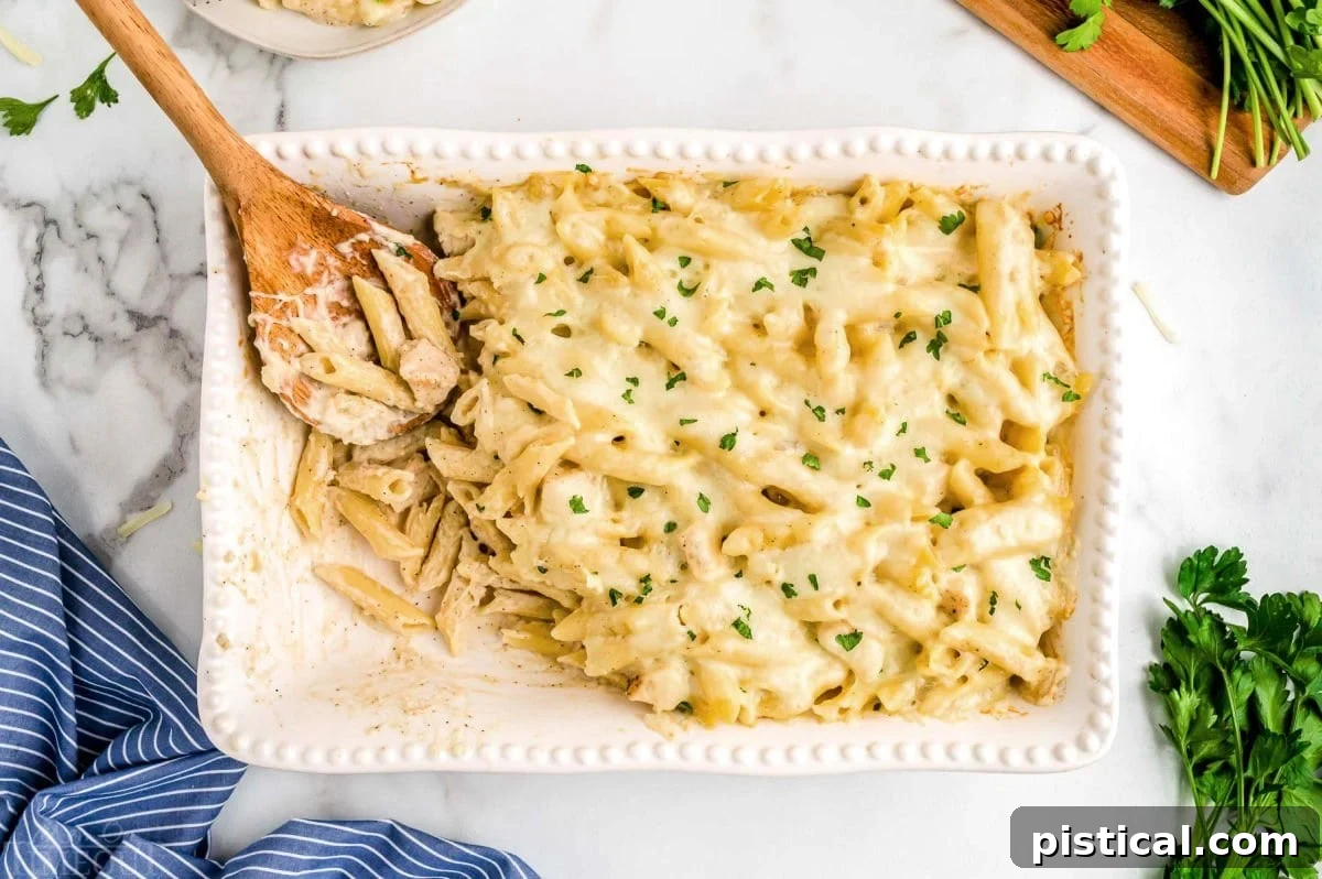 top down view of white casserole baking dish with some of the chicken alfredo bake already served. wood spoon is left in the dish.