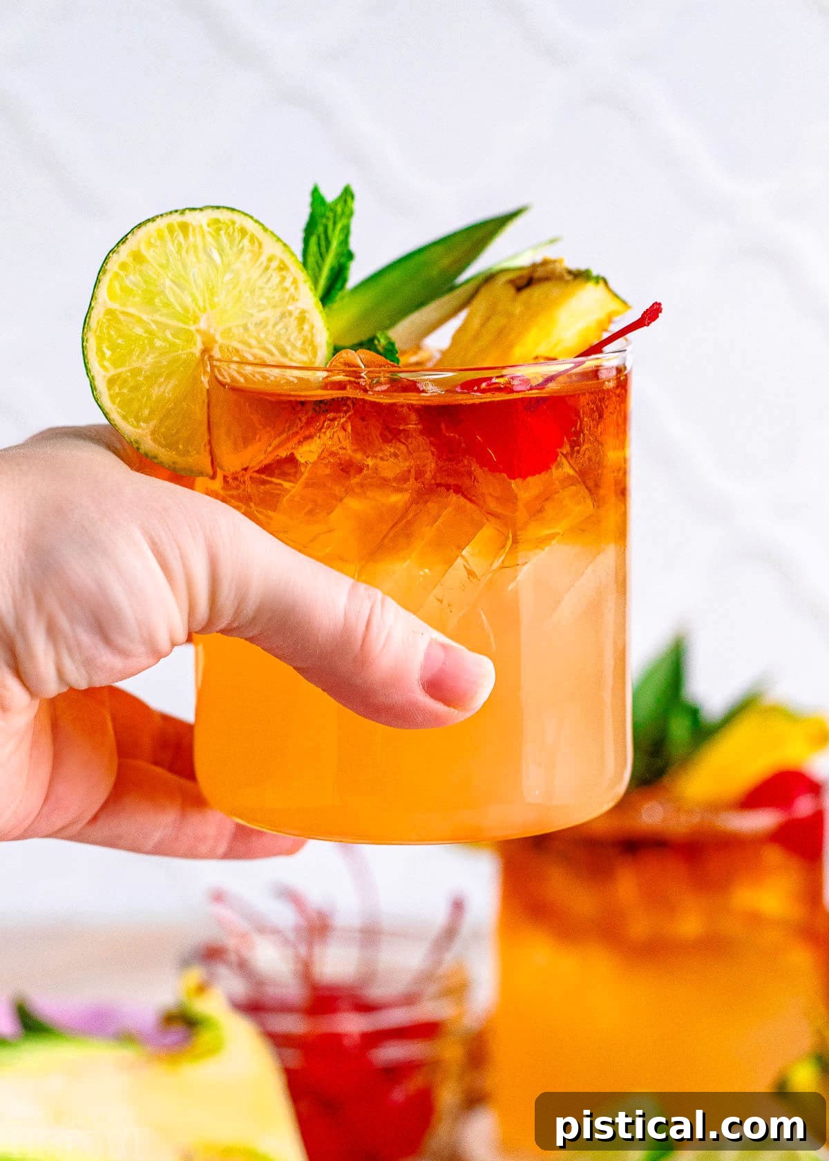 A Mai Tai cocktail being held up against a blurred background of a serving tray with another Mai Tai and liquor bottles, suggesting a vibrant cocktail setting.