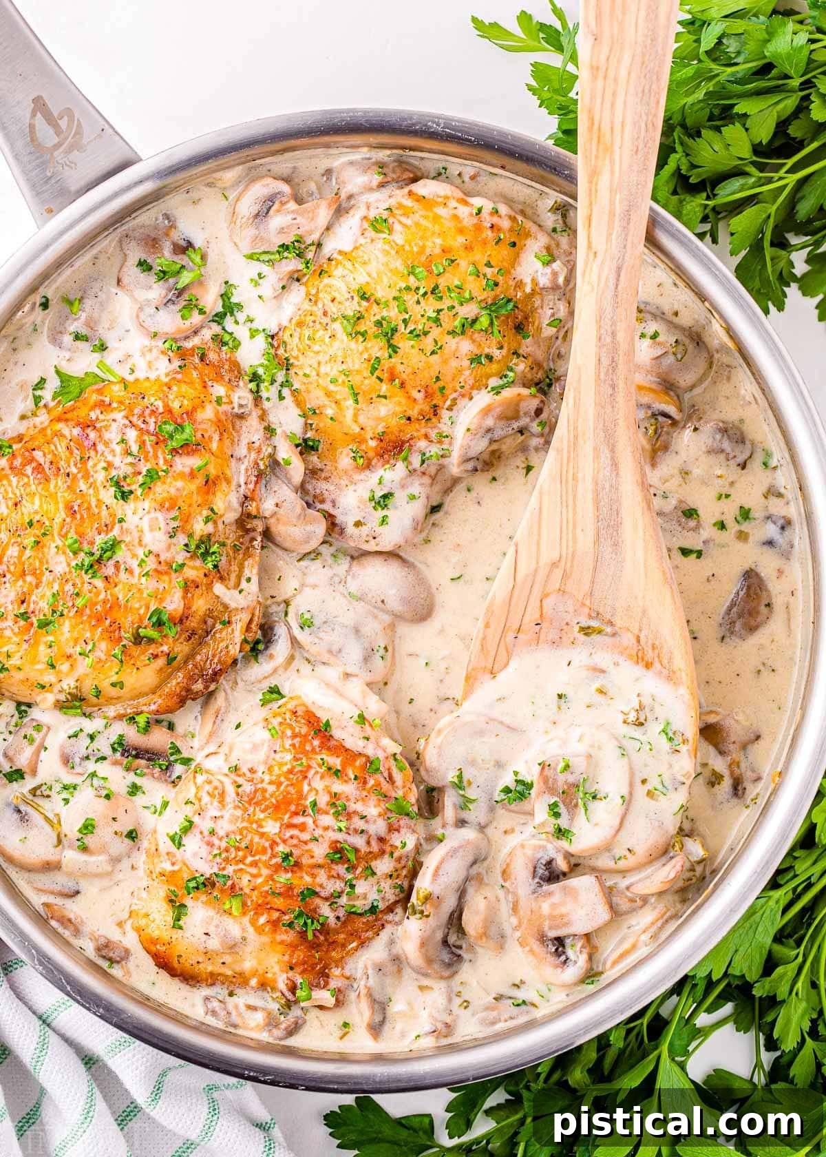 A top-down view of Chicken Fricassee simmering in a skillet, with a wooden spoon scooping out some of the creamy white wine mushroom sauce.