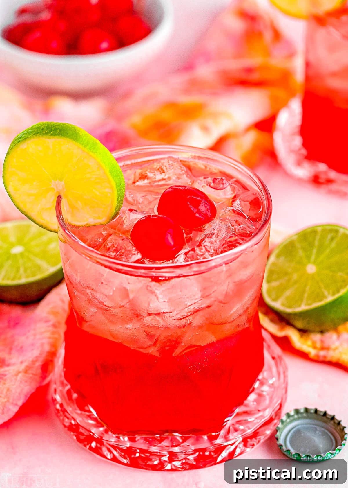 dirty shirley temple cocktail in a short glass garnished with a slice of lime and two maraschino cherries. Another drink and additional garnishes can be seen in the background.