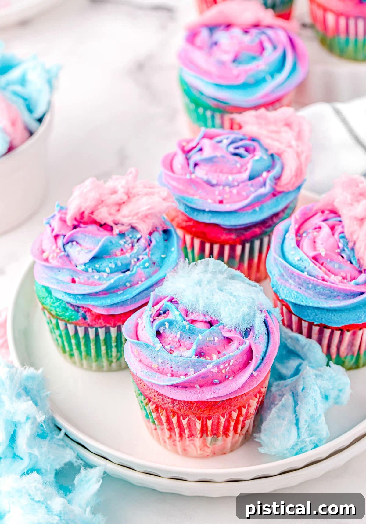 Five cotton candy cupcakes sitting on a white plate, topped with swirled pink and blue frosting and small pieces of cotton candy. Each cupcake is a burst of color and sweetness.