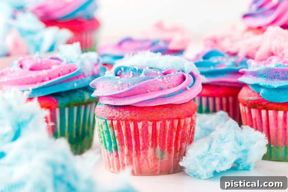 A collection of cotton candy cupcakes decorated with pink and blue frosting and topped with wisps of cotton candy, ready to be enjoyed. Scattered pieces of cotton candy surround the cupcakes.