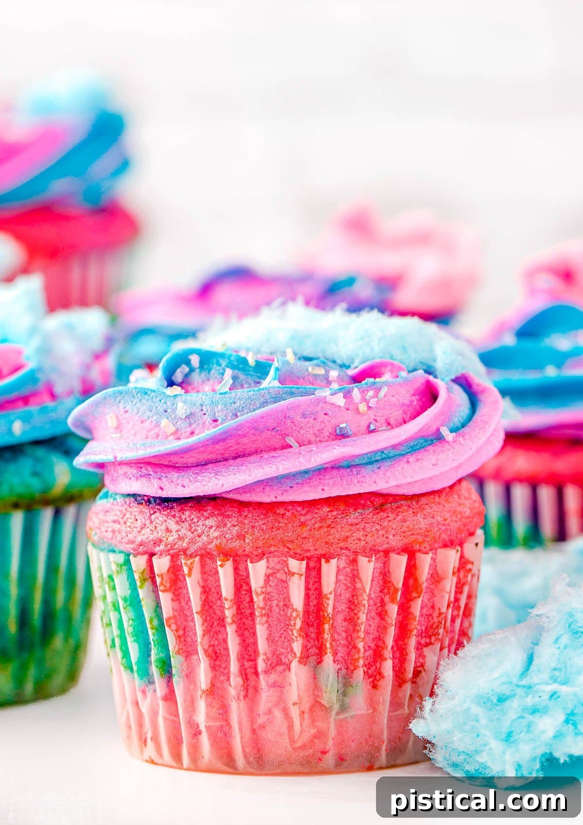 A display of numerous cotton candy cupcakes, decorated with both pink and blue frosting and adorned with cotton candy. Bits of cotton candy are sprinkled around the cupcakes for added visual appeal.