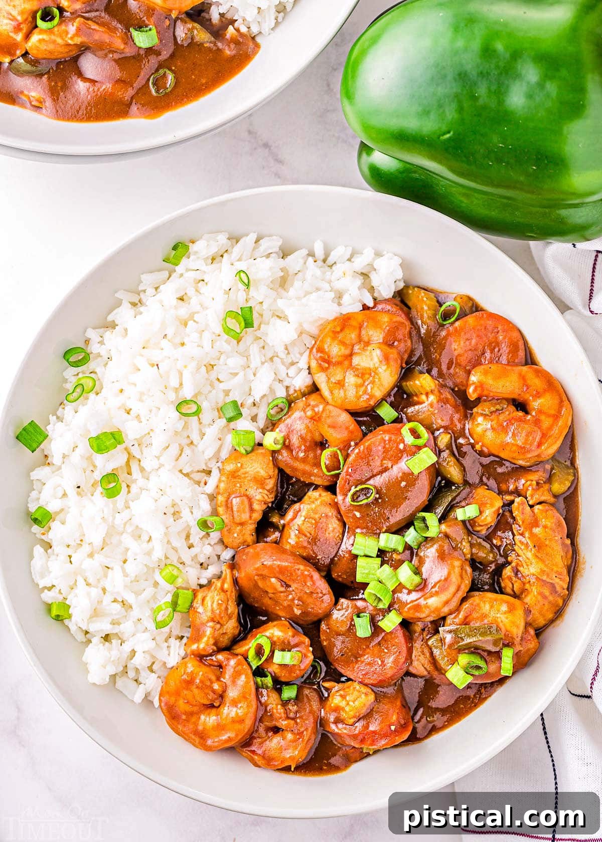 Foolproof Gumbo 6 A top-down view of a generously portioned bowl of seafood gumbo with white rice, garnished with vibrant green onions, positioned next to another plate and a green bell pepper.