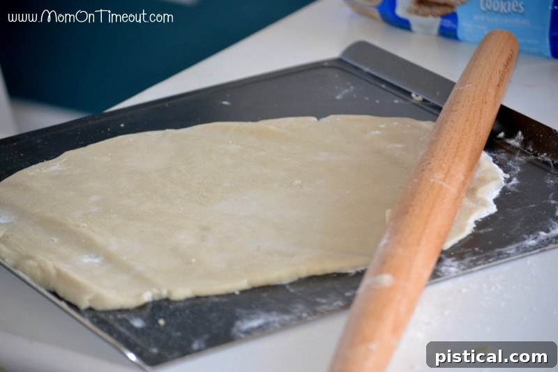 Prepping the base for Rainbow Sugar Cookie Bark: Rolling out refrigerated sugar cookie dough on parchment paper. Hands rolling out refrigerated sugar cookie dough on parchment paper