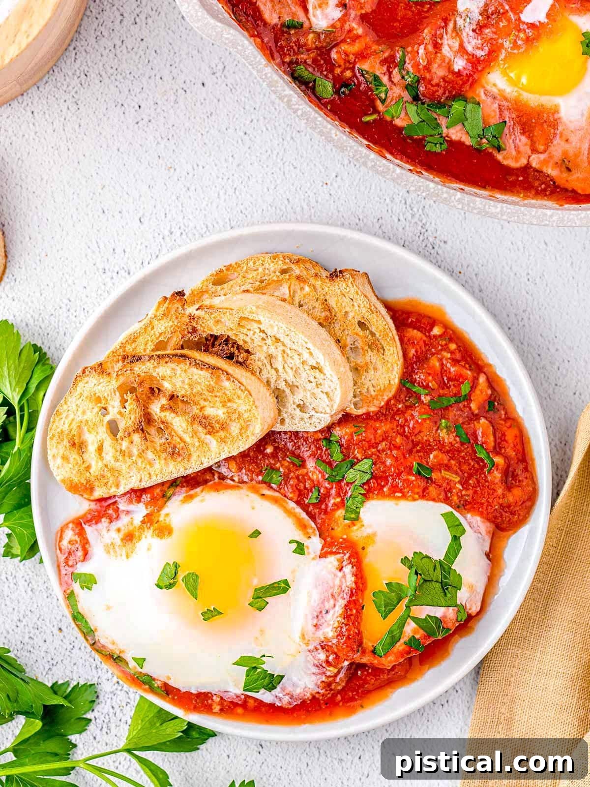 top down look at plated eggs in purgatory with two perfectly poached eggs rested in tomato sauce. Crusty, toasted bread on the plate. Garnished with chopped parsley.