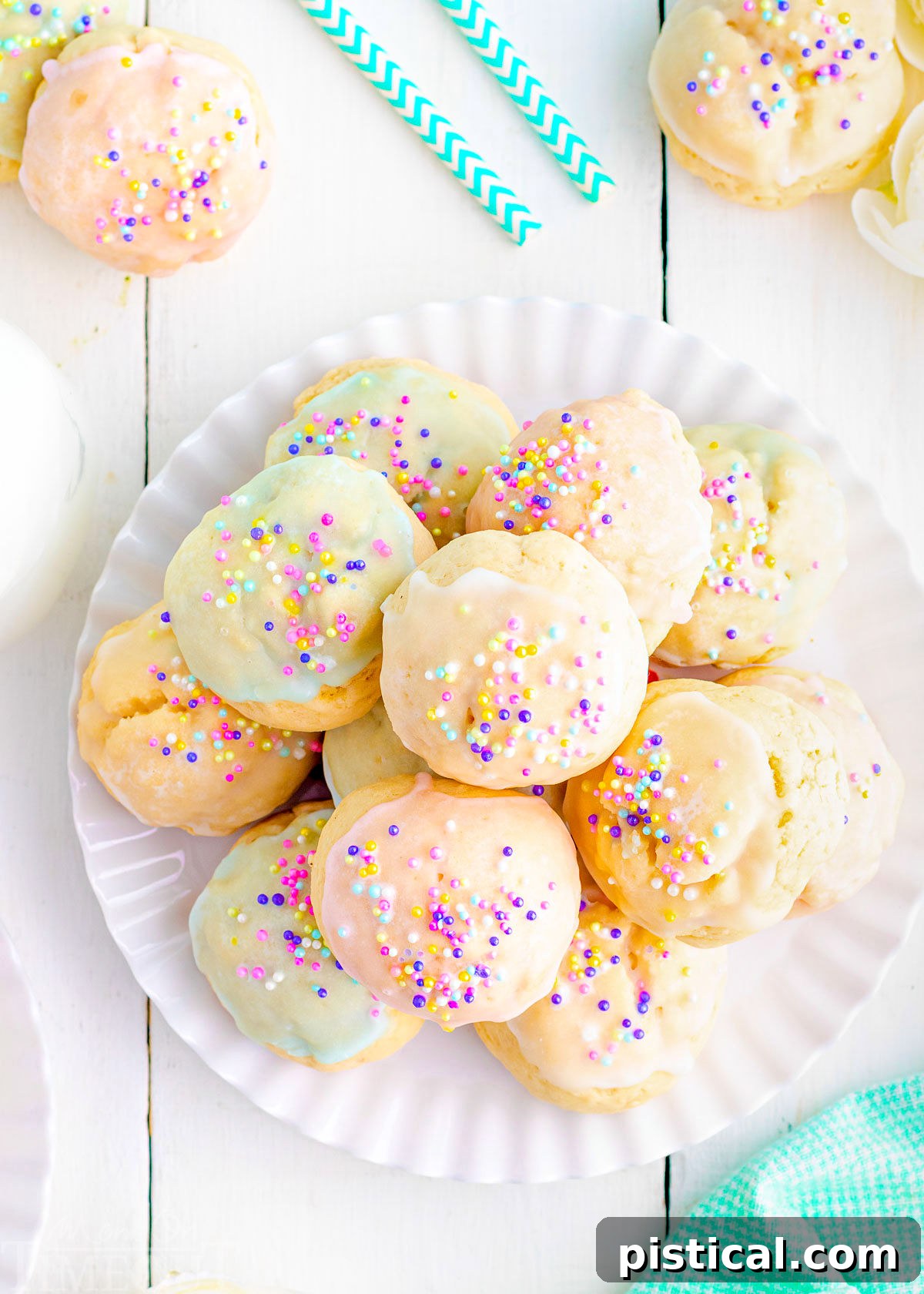 Top down view of a white plate piled high with cookies topped with pastel glazes and sprinkles. The plate is sitting on a white wood backdrop.