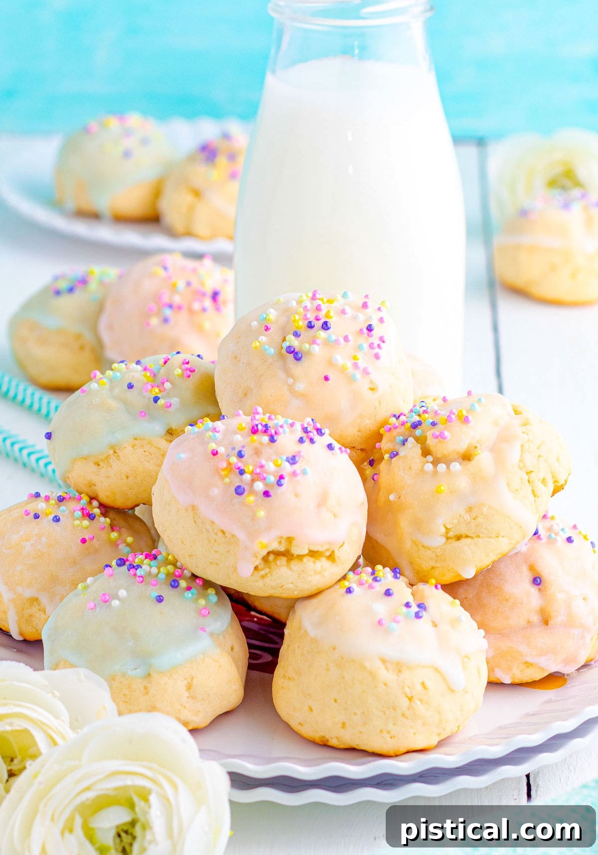 Plate of Italian Cookies sitting in front of a jug of milk and decorated with a pastel glaze and topped with sprinkles. Aqua background and more cookies can be seen in the background.