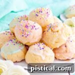 Wide view of colorful Italian cookies piled high on a white scalloped plate. Cookies are topped with sprinkles and pastel glaze in varying colors.