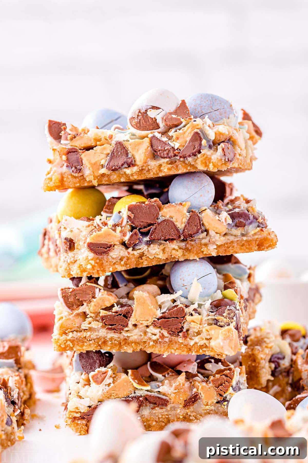 magic cookie bars topped with cadbury easter eggs stacked four high on a white surface. 