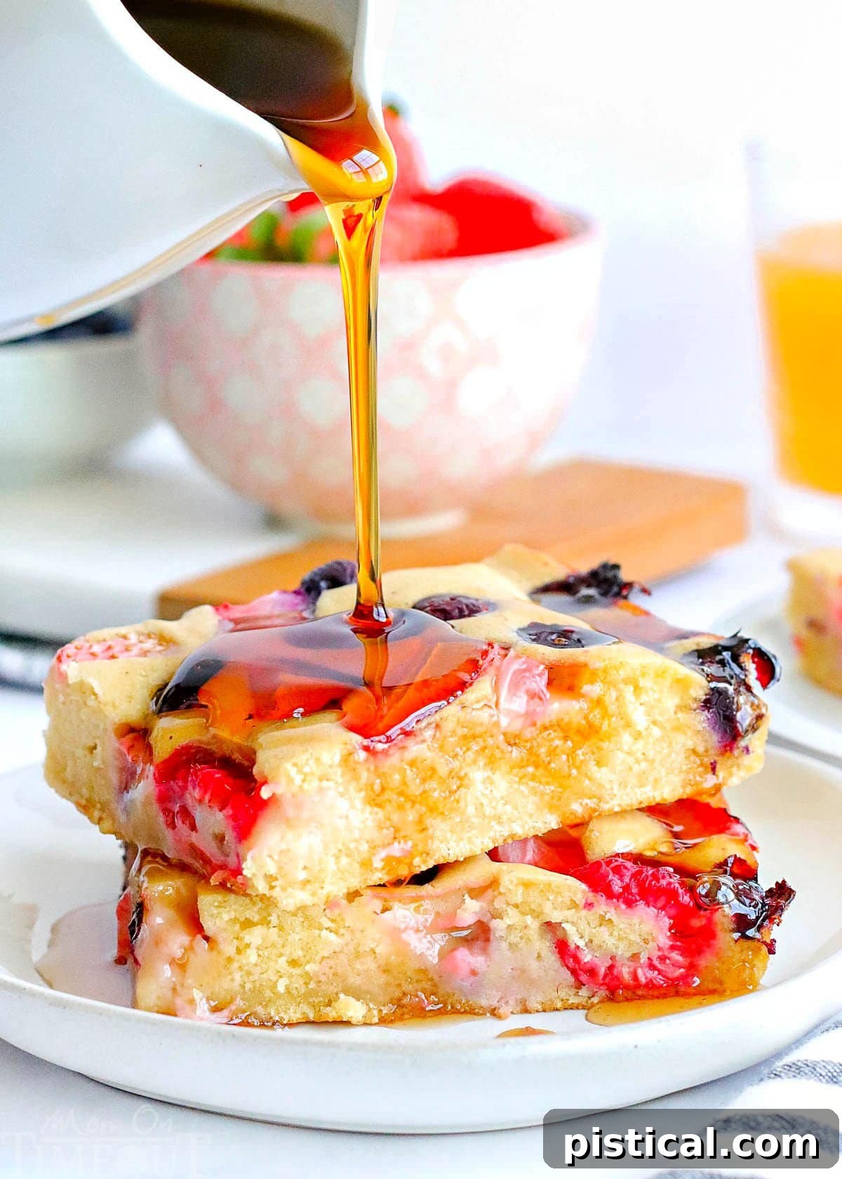 Two pieces of sheet pan pancakes made with three types of berries stacked on a white plate. Syrup is being poured on top of the pancakes, and a bowl of fresh berries can be seen in the background.