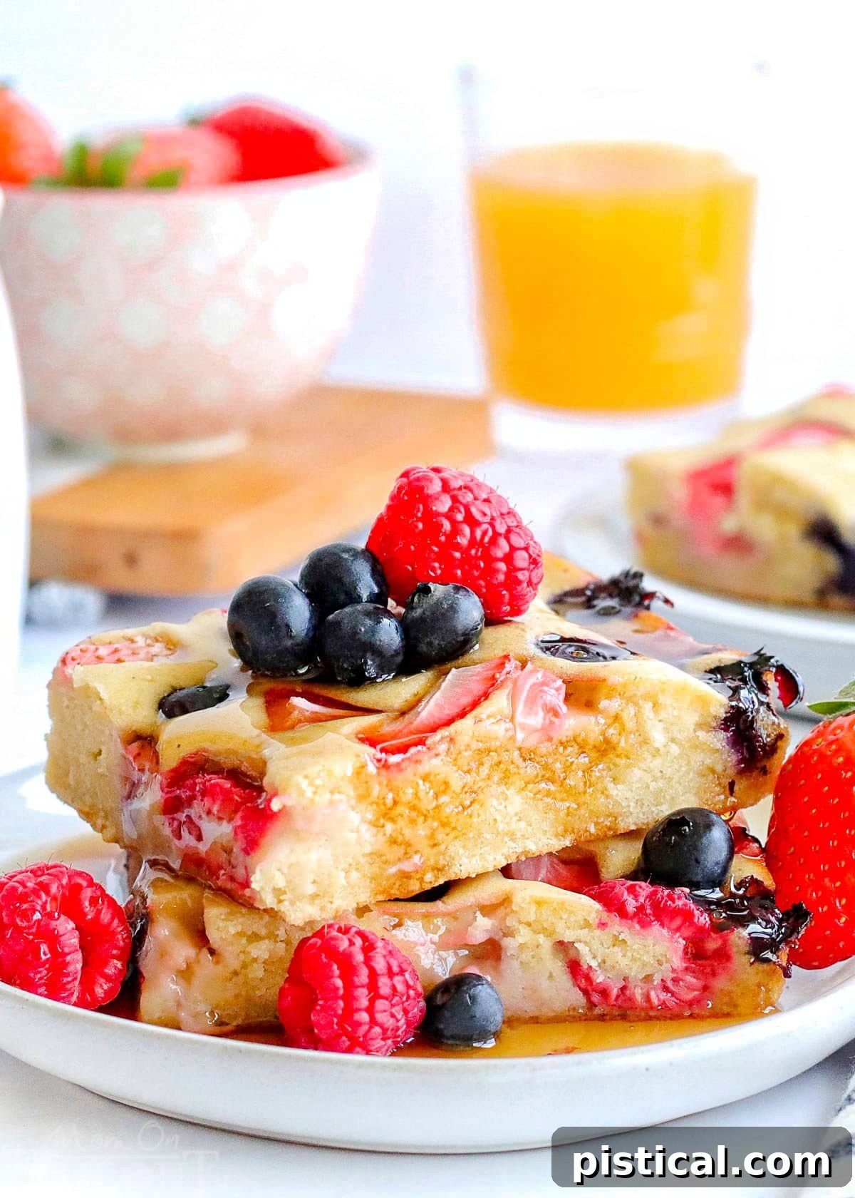 Two square-cut sheet pan pancakes, perfectly stacked on a white plate, are adorned with a vibrant assortment of fresh berries and a generous pour of golden maple syrup, ready to be enjoyed.