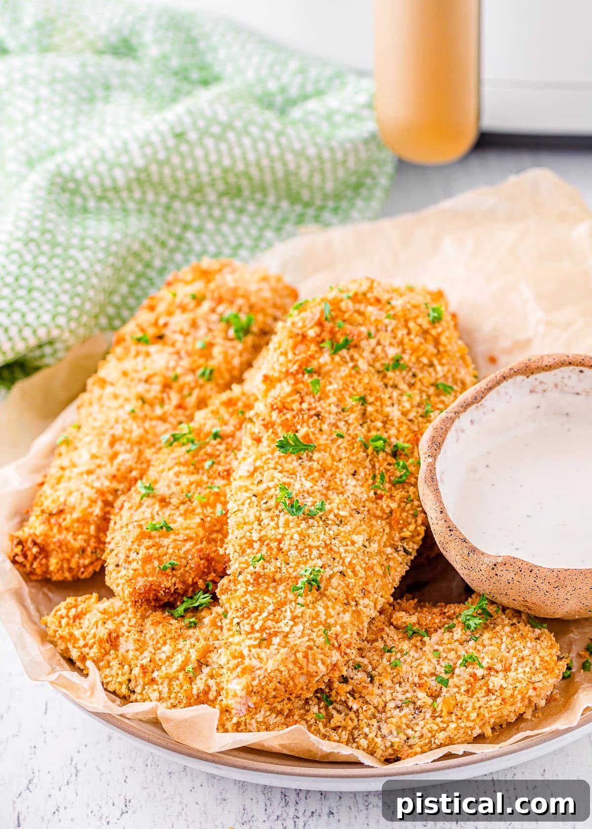 A close-up shot of cooked air fryer chicken tenders beautifully plated with a sprinkle of fresh parsley, and an air fryer in the background.