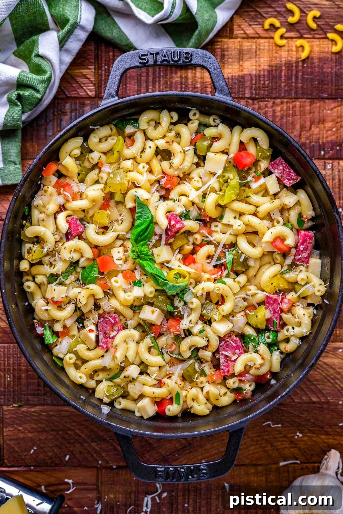 A beautiful top-down view of a generous portion of homemade Italian Pasta Salad, brimming with colorful ingredients and a zesty dressing, served in an elegant black round Staub dish with sturdy handles, resting on a rustic dark wood surface. This inviting image showcases the salad's readiness to be served and enjoyed at any gathering.
