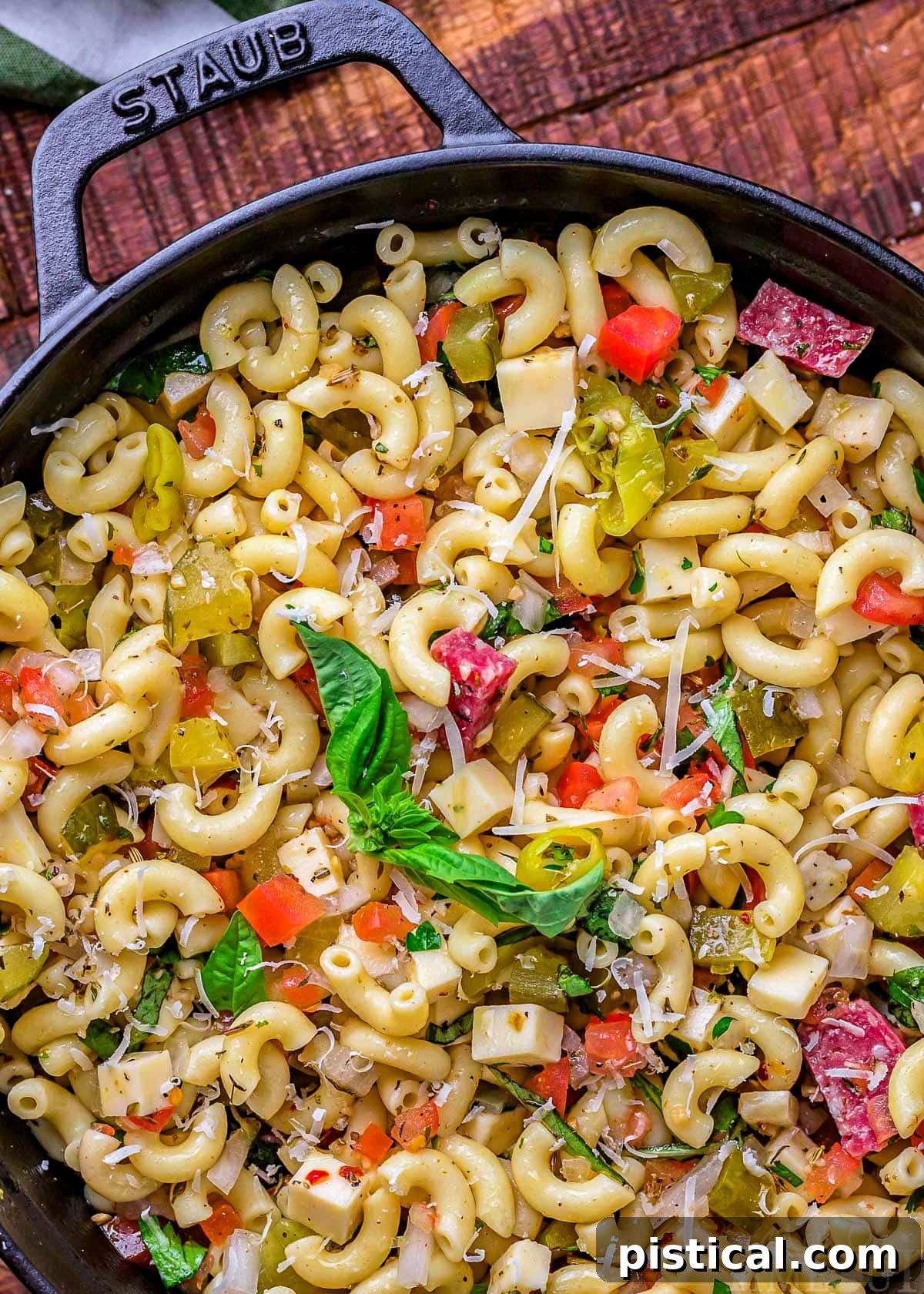 A close-up, appetizing view of a freshly prepared Italian pasta salad, nestled in a elegant black Staub casserole dish, ready to be served. The salad is beautifully garnished with vibrant green fresh basil leaves, highlighting its freshness and aromatic appeal.