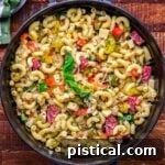 A captivating top-down view of a large black serving dish with handles, generously filled with Italian pasta salad tossed in a homemade Italian dressing. The vibrant pasta salad, featuring salami, pickles, and an array of fresh ingredients, is artfully arranged on a dark wood surface, emphasizing its rustic appeal and readiness for enjoyment.