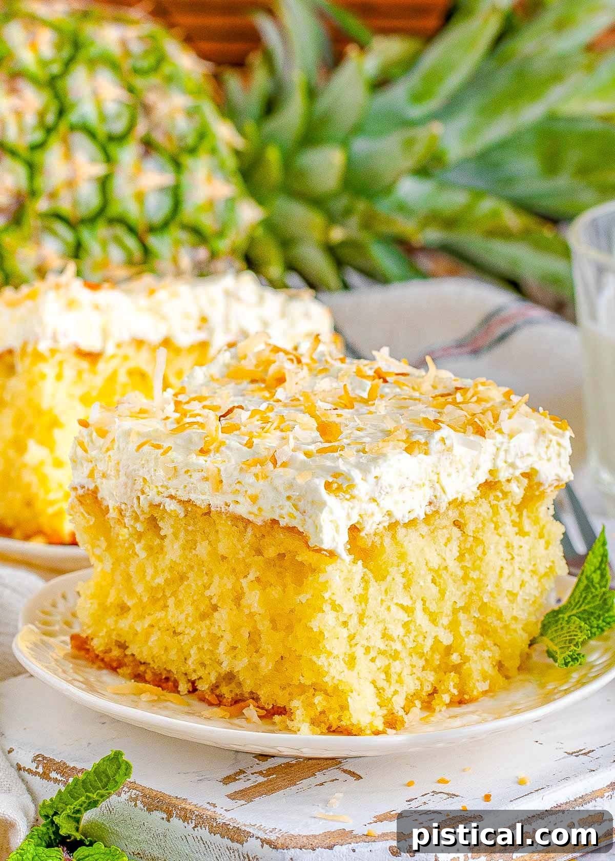 A square slice of moist Pina Colada Cake on a white plate, generously frosted with fluffy white coconut frosting and garnished with golden toasted coconut flakes. In the soft-focus background, another slice of cake and a fresh pineapple hint at the tropical theme.