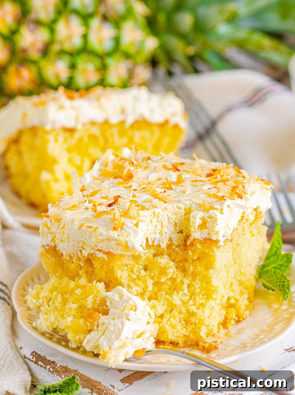 A tempting slice of Pina Colada Poke Cake resting on a white plate, with a fork gently pulling away a piece, highlighting its moist interior and luscious frosting. Another slice and a fresh pineapple are visible in the background, reinforcing the tropical theme.