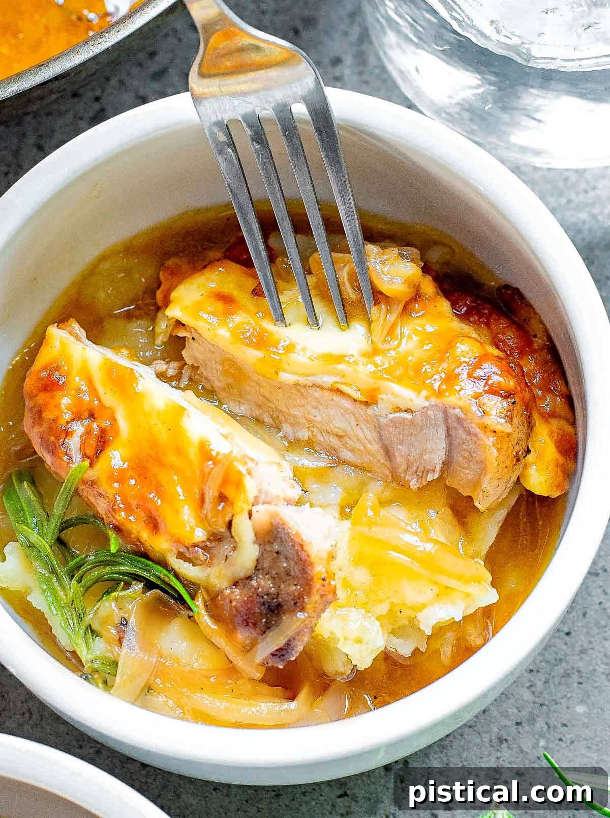pork chop in small bowl on top of mashed potatoes cut in half so the juicy interior is showing. a fork is holding the two pieces apart.