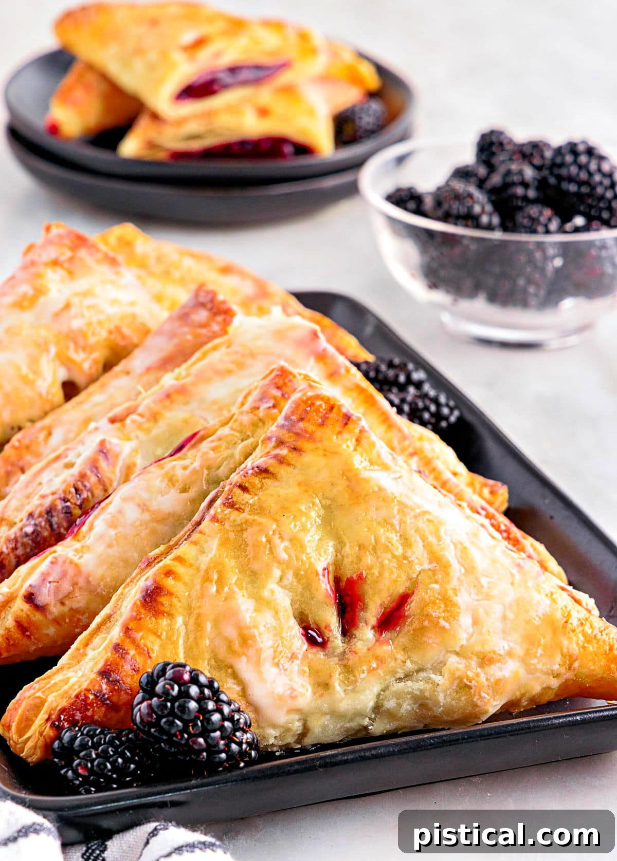A row of golden-brown blackberry turnovers neatly lined on a black rectangular plate, with a bowl of fresh blackberries nearby.