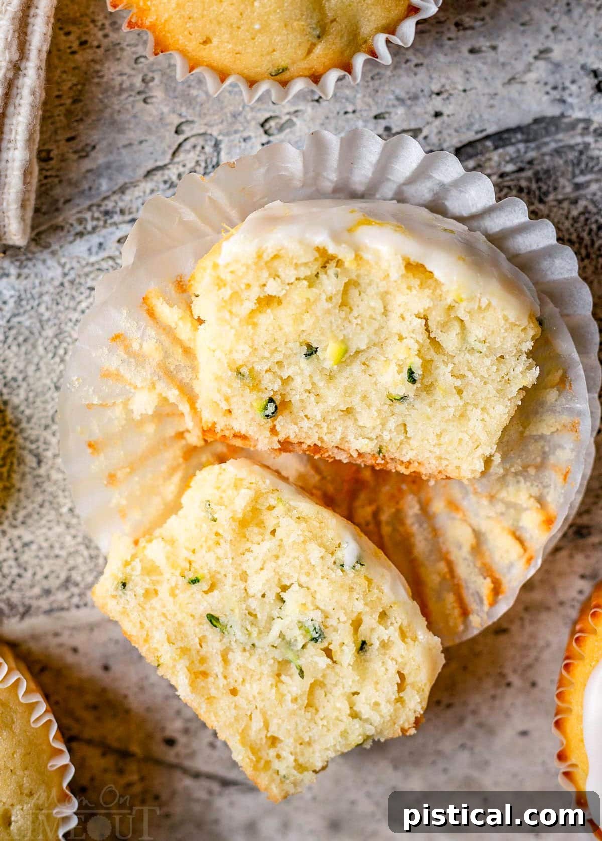 Zesty Lemon Zucchini Muffins 2 lemon zucchini muffin in white liner topped with lemon glaze cut in half and sitting on a gray backdrop.