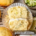 lemon zucchini muffin cut in half with one half resting on the other. three whole muffins and some shredded zucchini are near the cut muffin.