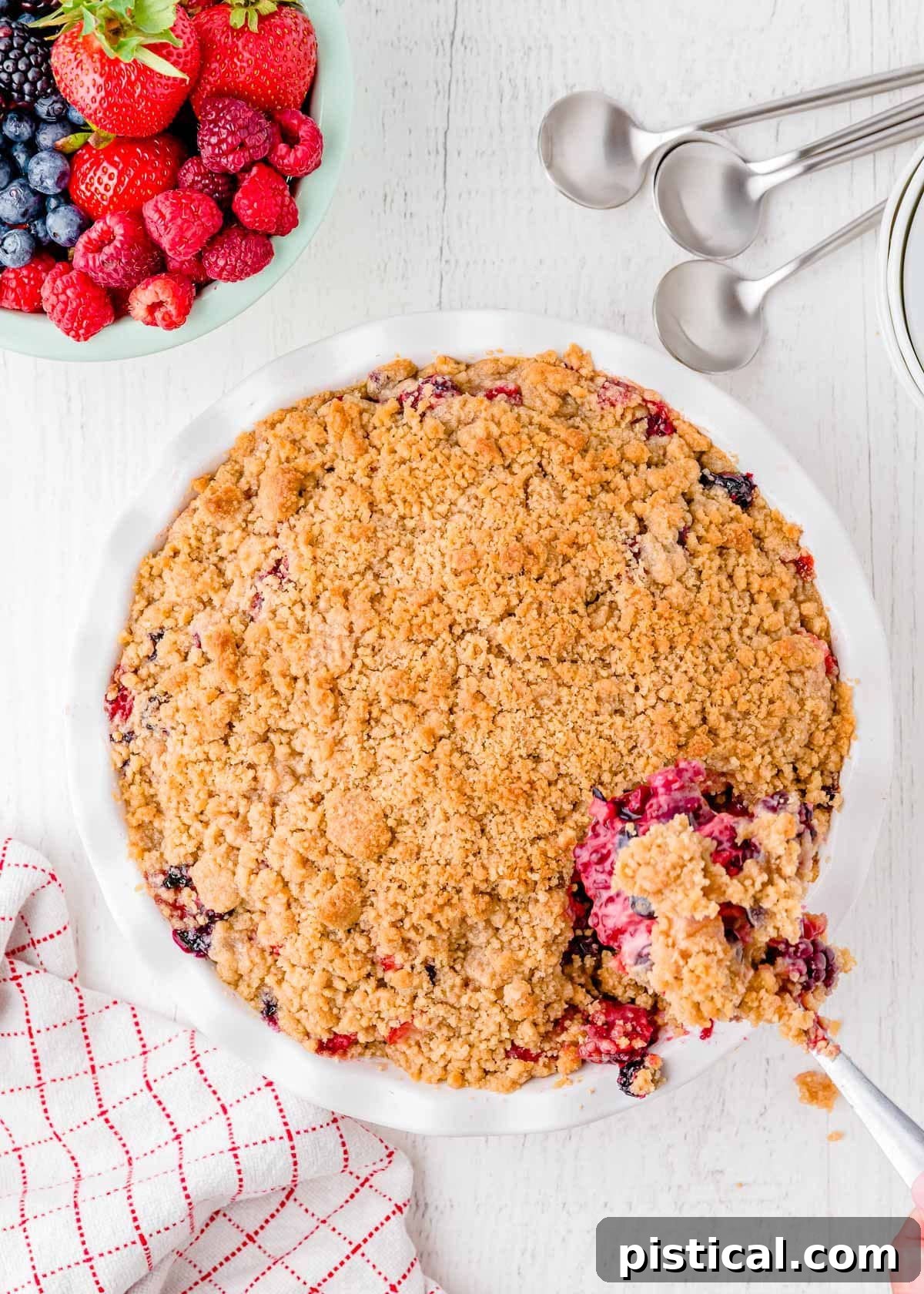 A beautiful top-down perspective of a golden-brown berry crumble, baked in a rustic white pie dish, with a serving spoon gently lifting a portion to reveal the rich, bubbling fruit filling.