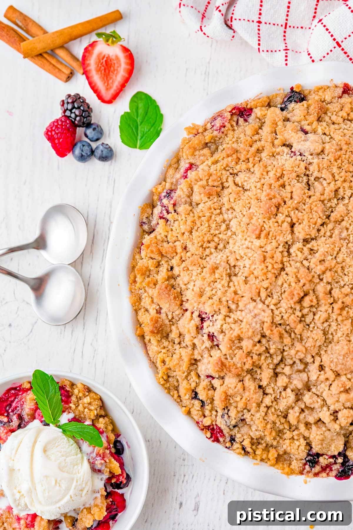 A top-down view of a golden-brown berry crumble, baked in a white pie plate, with a single perfect serving scooped out and placed in an accompanying white bowl. Both the main dish and the individual serving are lavishly topped with melting vanilla ice cream, creating an irresistible visual.