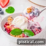 A white bowl with warm berry crumble, topped with vanilla ice cream. A spoonful is being scooped out, showcasing the dessert's delicious texture.