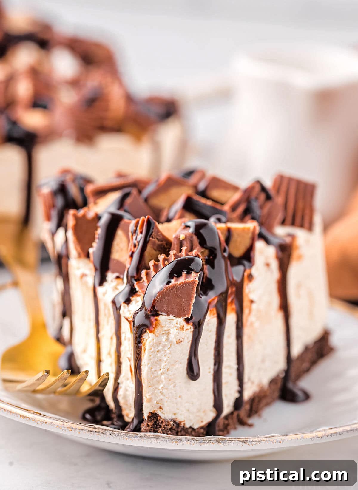 A delectable slice of no-bake peanut butter cheesecake, adorned with Reese's candies and a generous pour of hot fudge, rests on a small gold-rimmed white plate with a golden fork beside it.