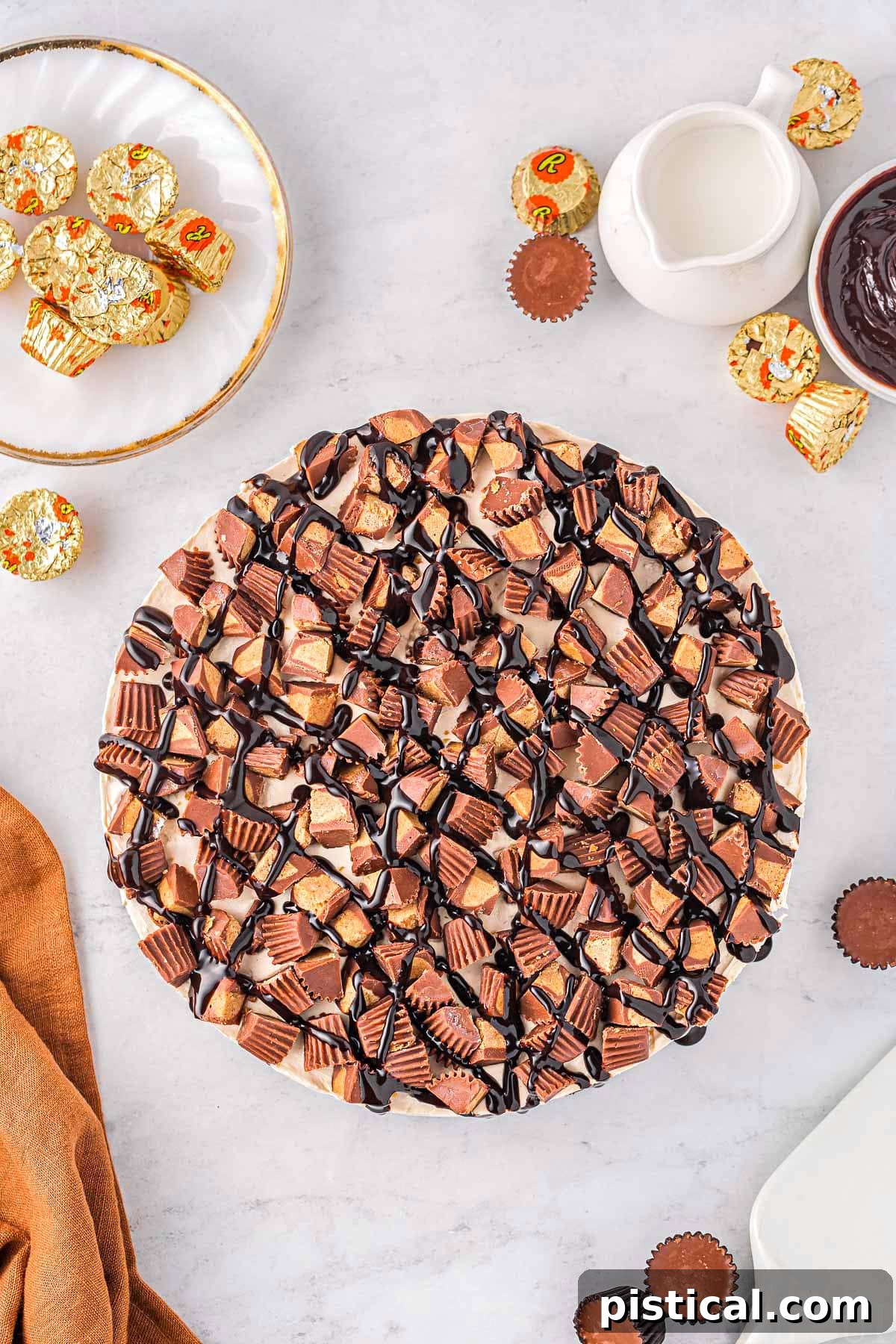 A top-down view of a whole peanut butter cheesecake, generously topped with Reese's candies and hot fudge, ready to be sliced and enjoyed.