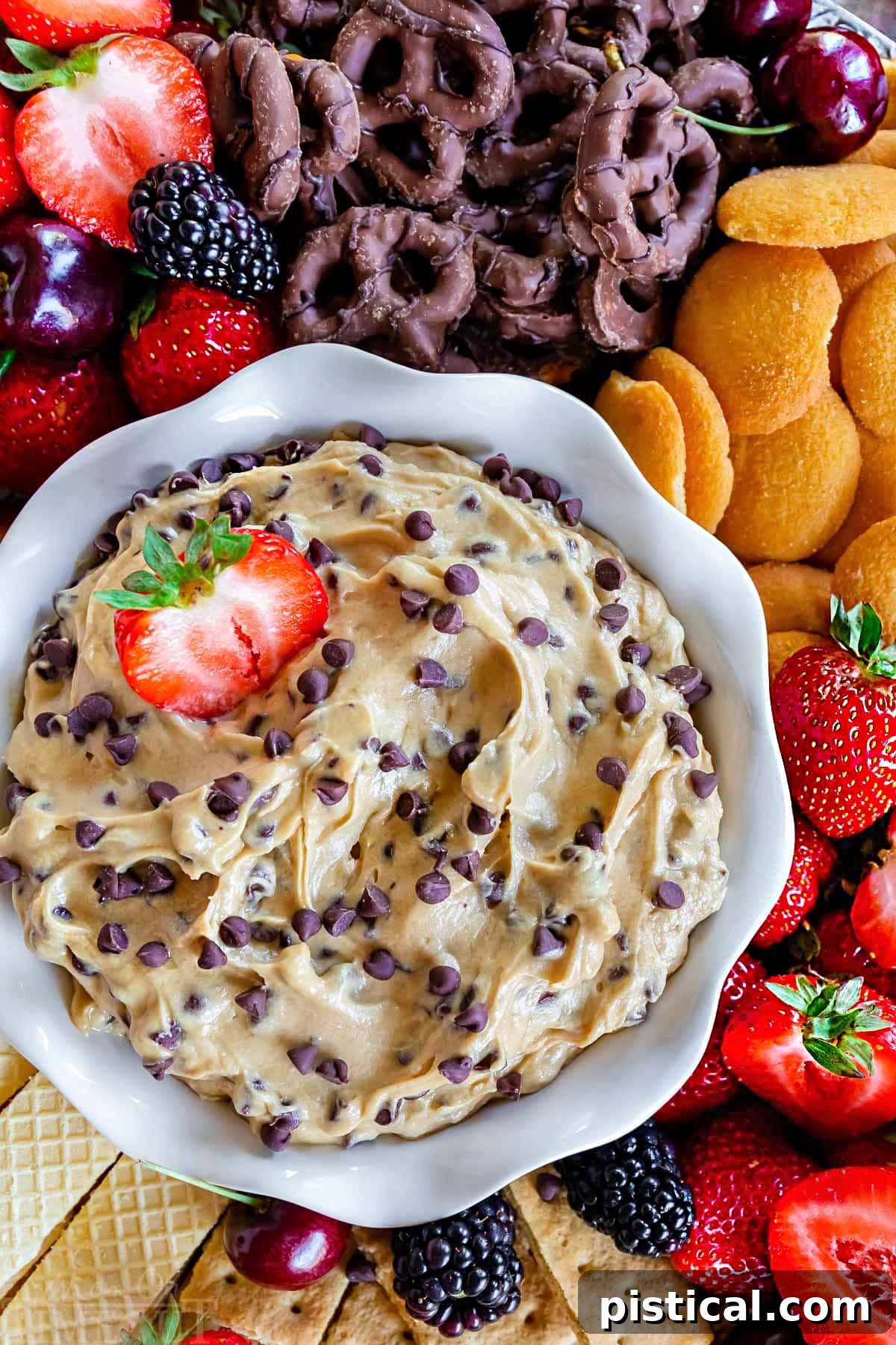 A top-down view of a white bowl filled with creamy chocolate chip cookie dough dip, elegantly surrounded by an assortment of dippers including vibrant strawberries, decadent chocolate-dipped pretzels, classic Nilla wafers, and other fresh fruits and cookies. This image highlights the dip's irresistible texture and the variety of serving possibilities.