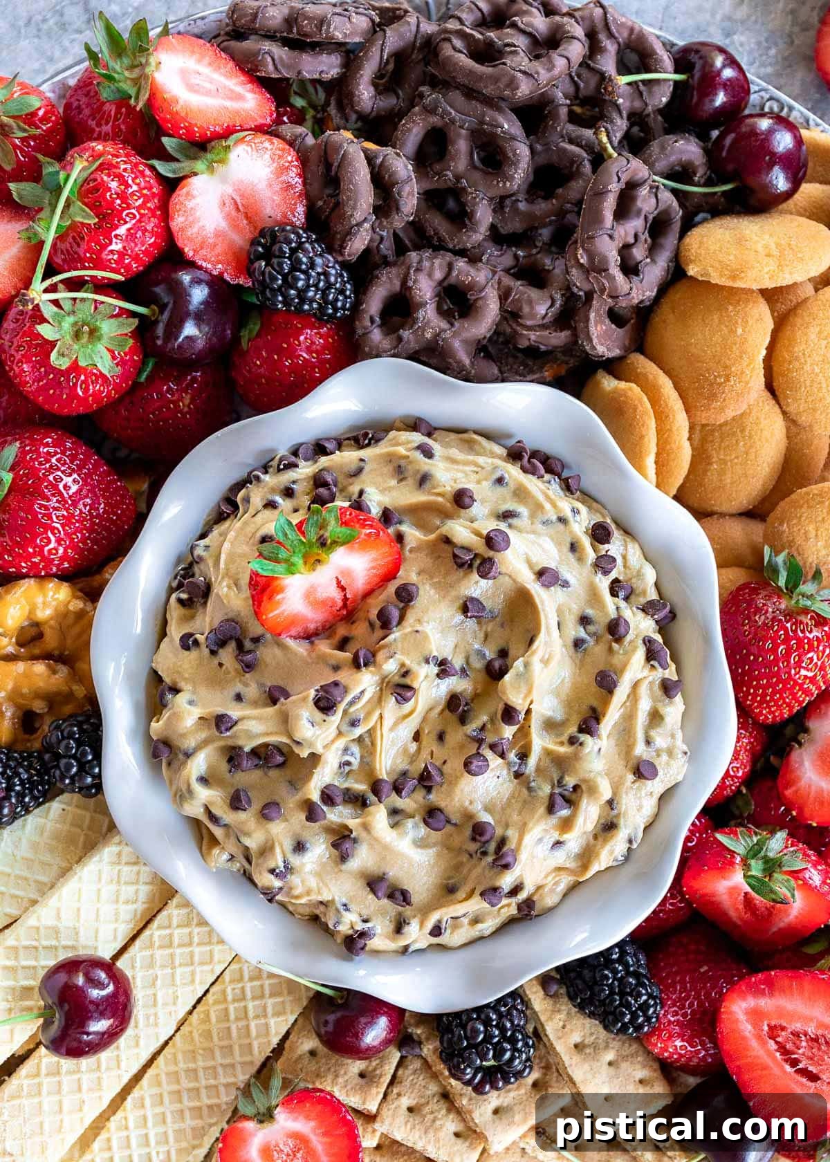 A top-down perspective of a pristine white bowl generously filled with delectable chocolate chip cookie dough dip. The bowl is artfully arranged with a tempting array of dippers, including succulent strawberries, crispy chocolate-dipped pretzels, golden Nilla wafers, and other vibrant fruits and cookies, highlighting the dip's versatility.