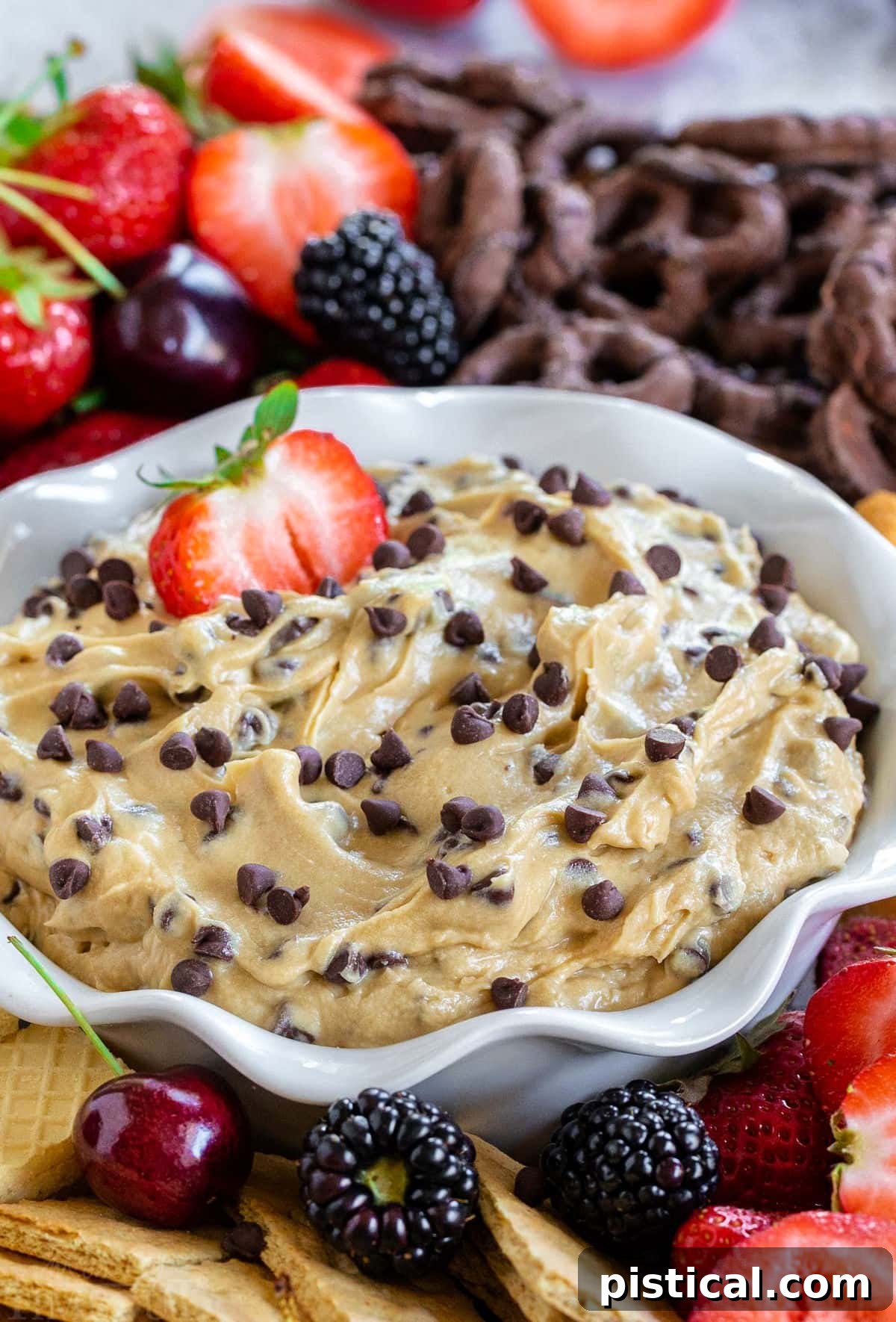 A charming bowl with elegant fluted edges holds a generous portion of creamy chocolate chip cookie dough dip, with a glistening half-strawberry perfectly dipped in. The bowl is beautifully adorned with a vibrant assortment of fresh fruit, including strawberries and blueberries, and an array of tempting cookies, inviting guests to enjoy.