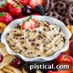 A bowl with fluted edges holds chocolate chip cookie dough dip with half a strawberry dipped into the dip. Fresh fruit and cookies surround the bowl.