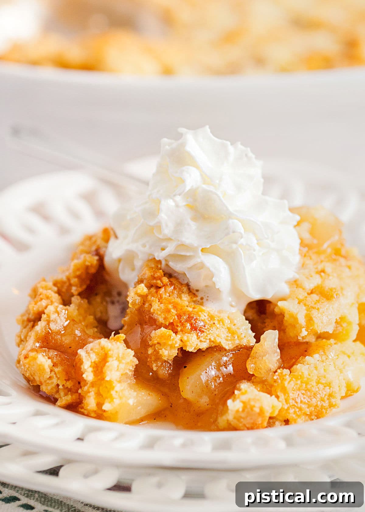 Golden Apple Cobbler Delight 2 scoop of apple cobbler served on a white lace edged plate topped with fresh whipped cream. white casserole dish with remaining cobbler in the background.