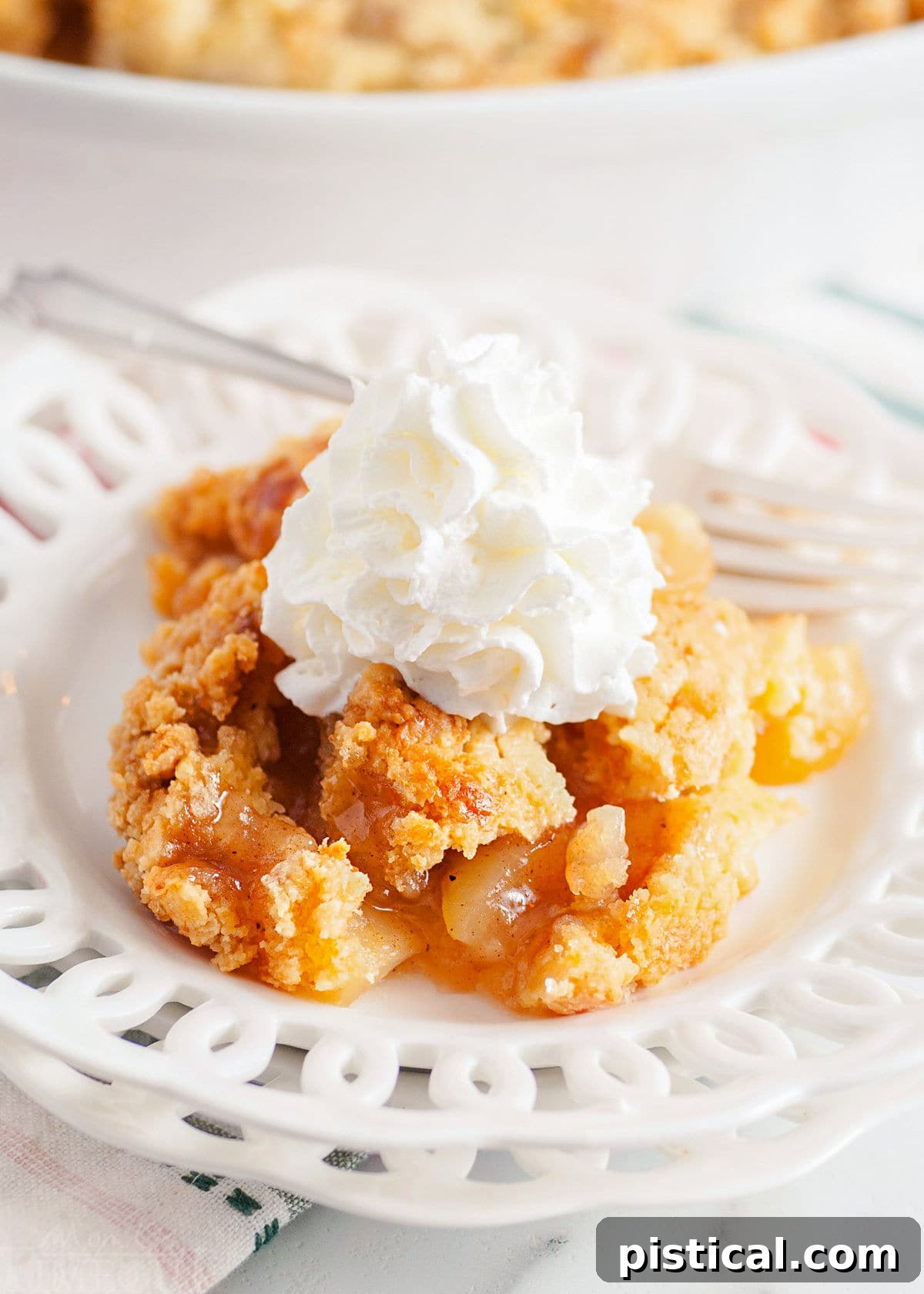Golden Apple Cobbler Delight 8 scoop of apple cobbler served on a white lace edged plate topped with fresh whipped cream. white casserole dish with remaining cobbler in the background.