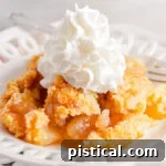 scoop of apple cobbler served on a white lace edged plate topped with fresh whipped cream. white casserole dish with remaining cobbler in the background.