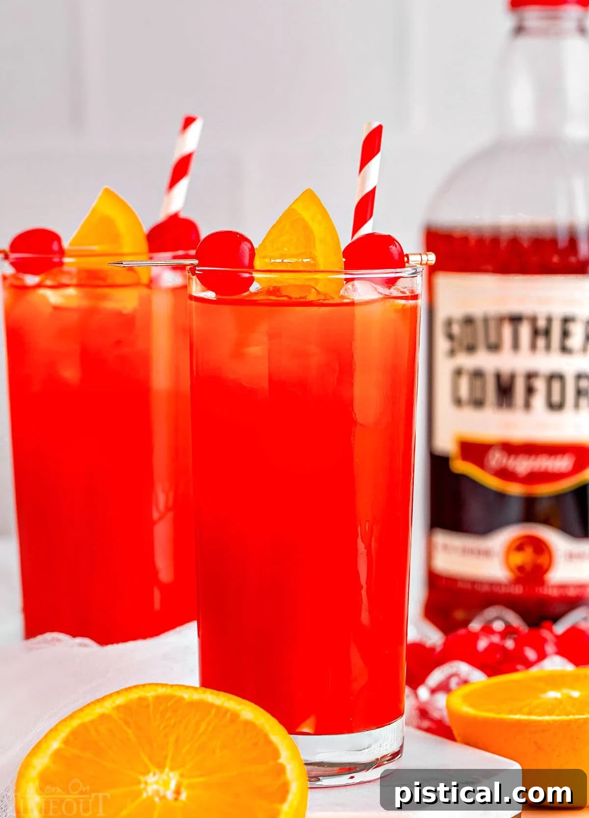 Two tall glasses filled with a vibrant Alabama Slammer cocktail, adorned with red and white striped straws. A bottle of Southern Comfort and a halved orange are visible in the soft-focus background.