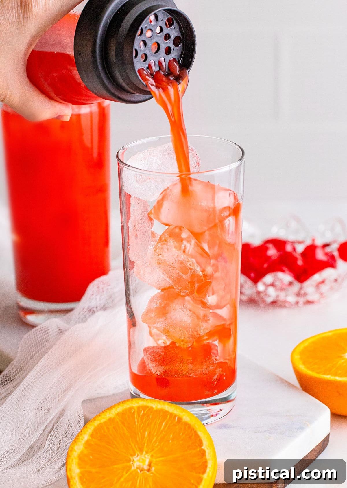 An Alabama Slammer cocktail being skillfully poured from a shaker into a tall glass filled with ice, with another finished drink visible in the background.