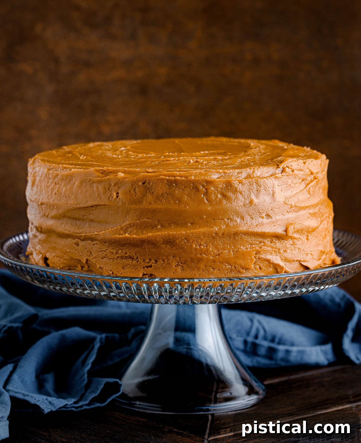 A beautifully frosted two-layer caramel cake sits elegantly on a glass cake stand, with a soft navy blue napkin providing a gentle backdrop. This homemade caramel cake is a perfect dessert for any occasion.