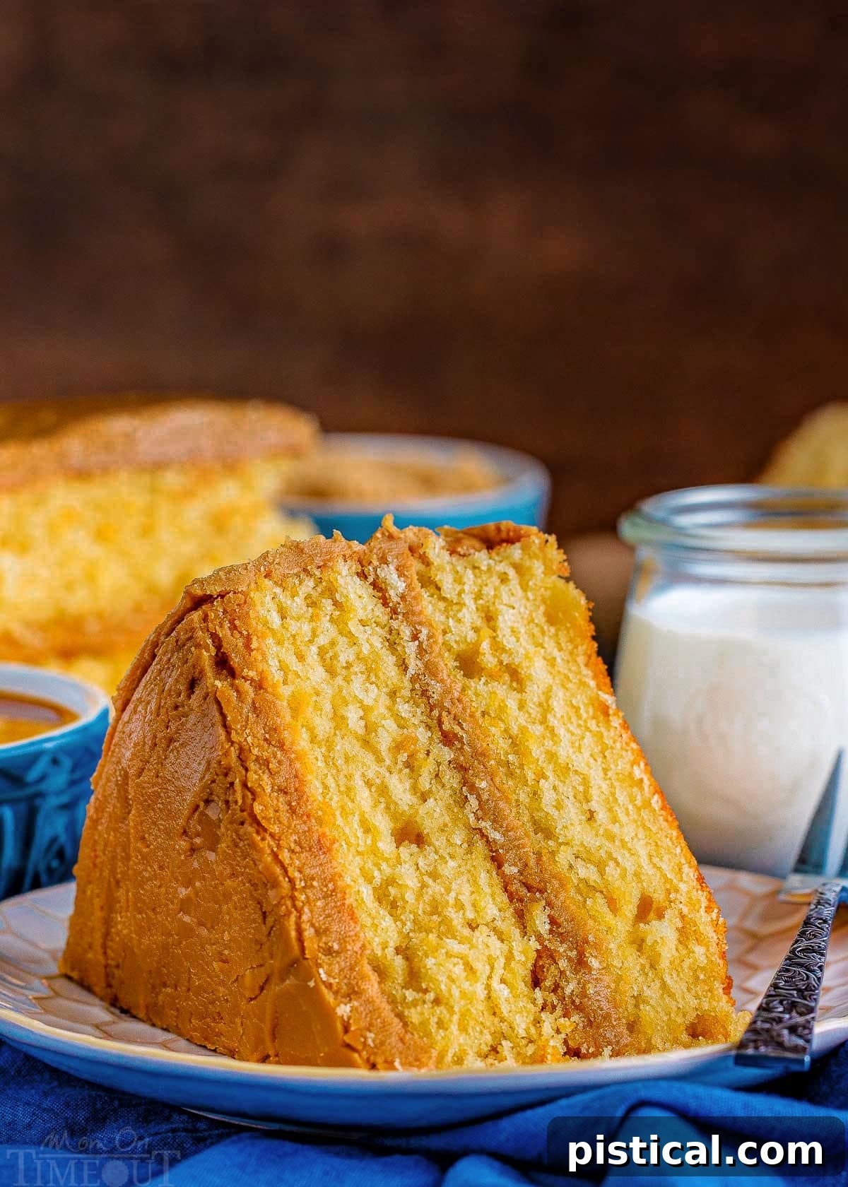 A perfectly sliced piece of caramel layer cake rests on a white plate, showcasing its moist layers and rich caramel frosting. Another slice and a small glass of milk are in the background, inviting indulgence.