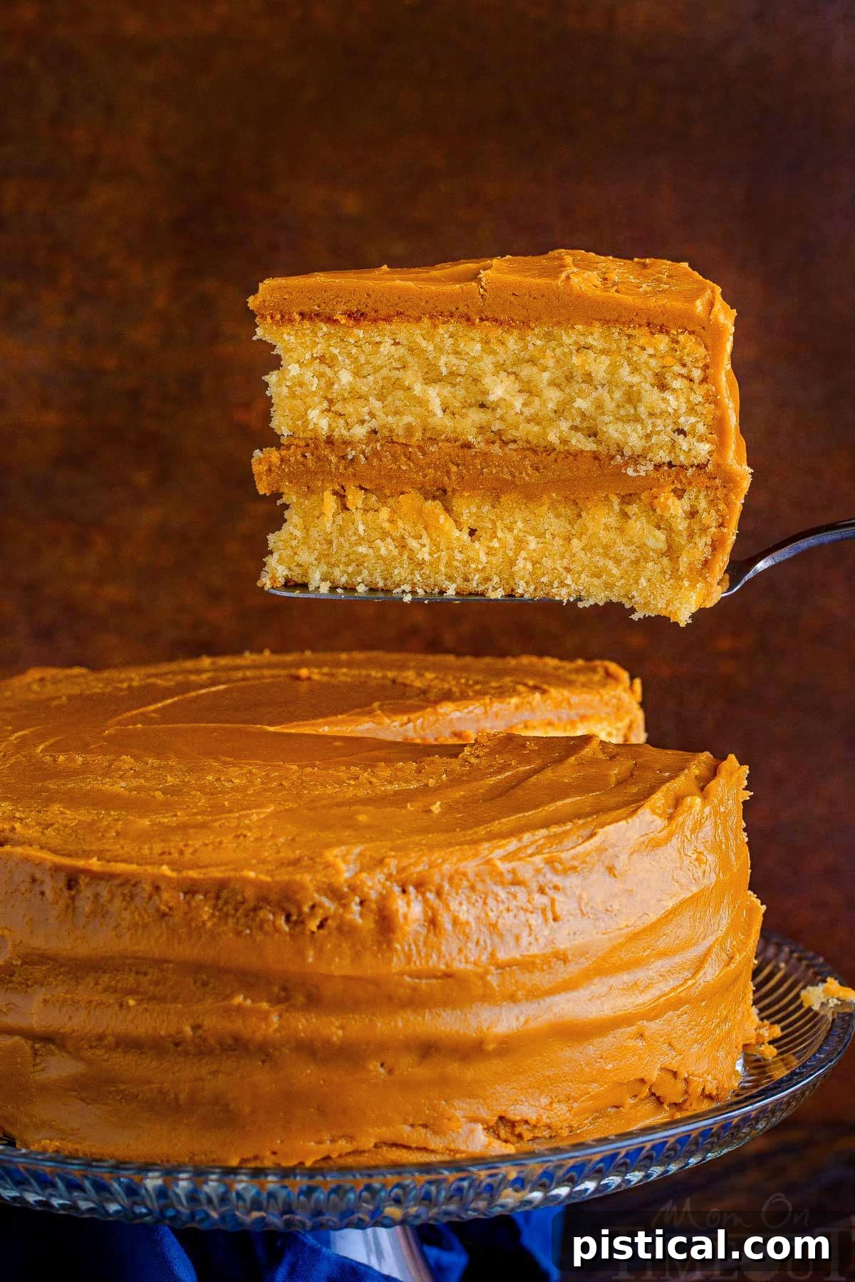 A hand holds a perfect slice of caramel cake, lifted slightly above the remaining cake on a cake stand, highlighting the rich frosting and tender layers.