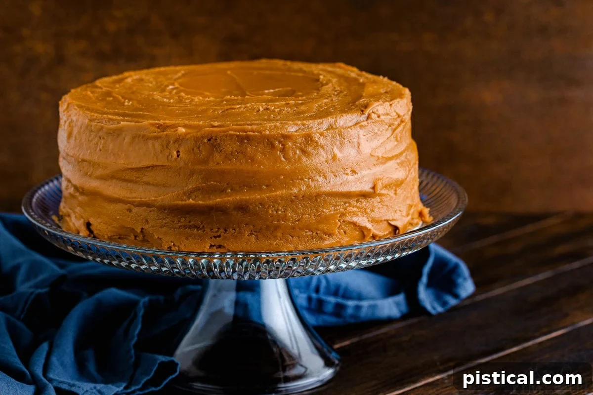 A magnificent two-layer caramel cake, generously covered in luscious caramel frosting, rests on a gleaming glass cake stand, with a tasteful navy blue napkin providing a serene background. This easy caramel cake recipe is perfect for any occasion.