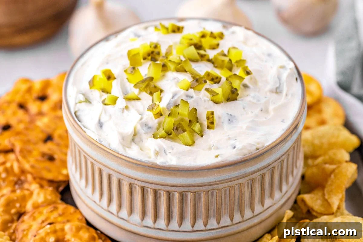 Dillicious Dip 4 A small, decorative gray clay bowl with ridged details, overflowing with dill pickle dip and garnished with extra diced pickles. An assortment of pretzels and chips surrounds the bowl, ready for dipping.