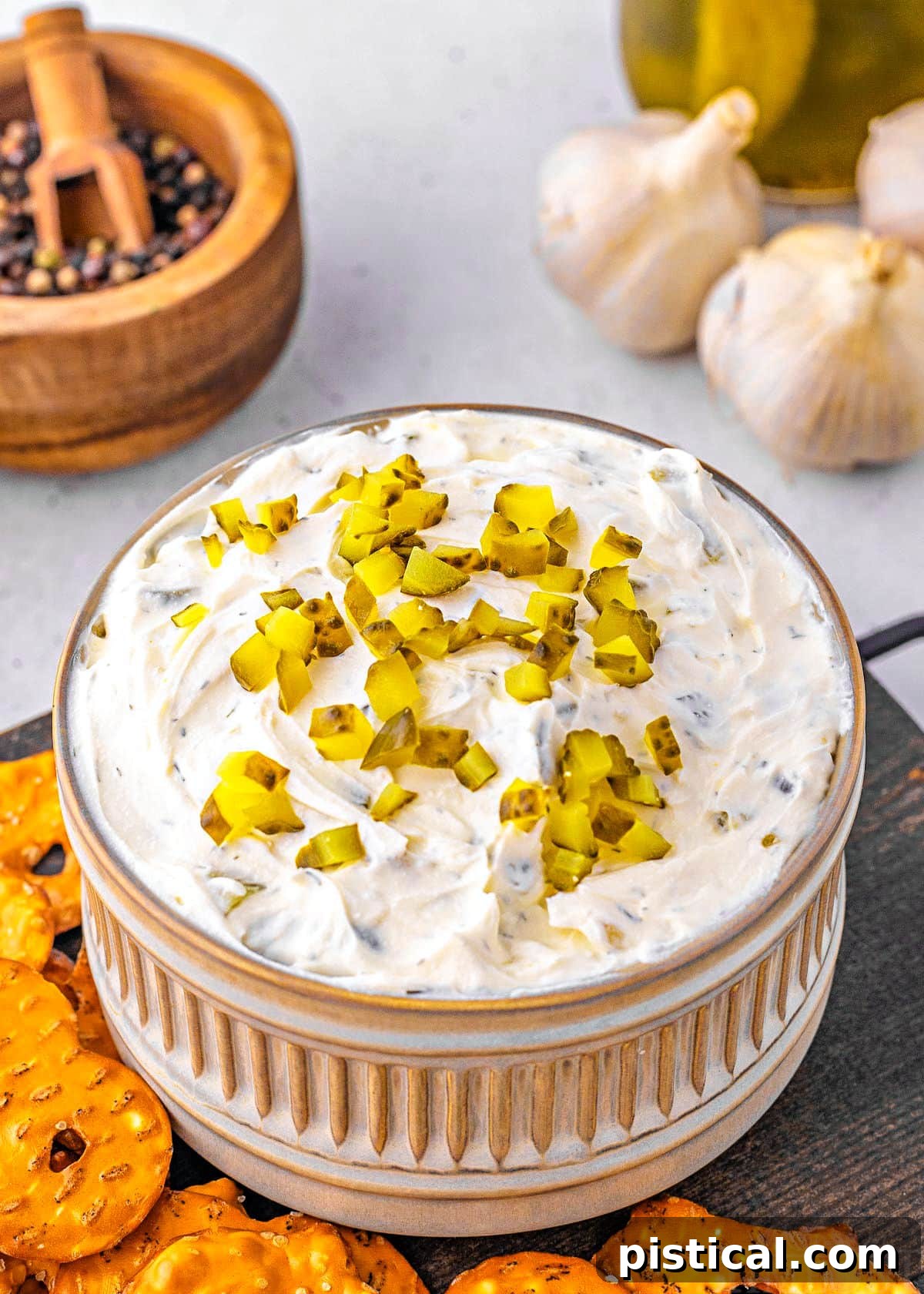 Dillicious Dip 6 A small, elegant gray clay bowl, textured with ridges, holds a generous amount of dill pickle dip, topped with more finely diced pickles. The bowl is artfully surrounded by a selection of crunchy pretzels and chips, inviting guests to dip.